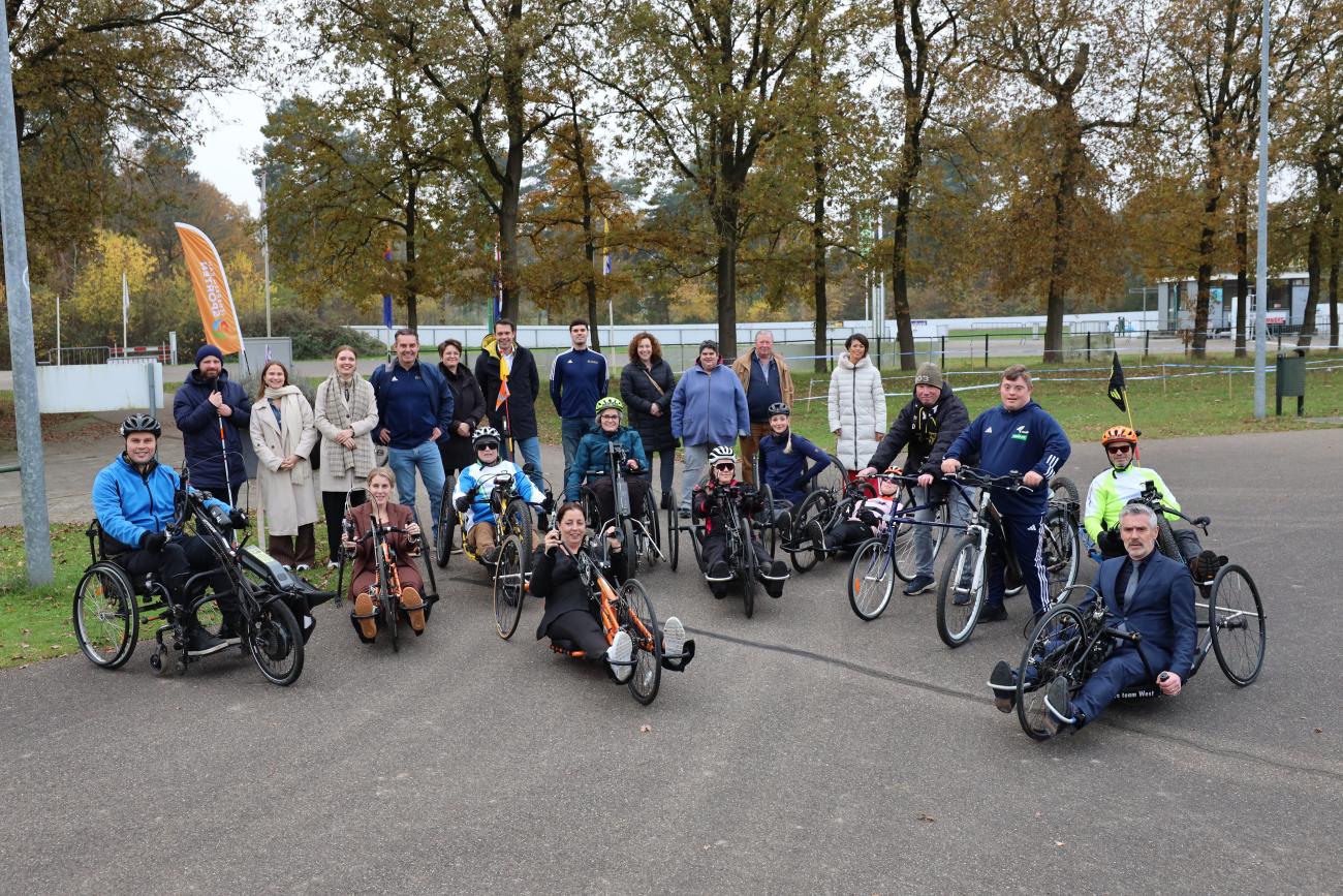 Een groepsfoto van alle betrokkenen bij het werkbezoek van staatssecretaris Maeijer, waarbij ze op een handbike zitten.