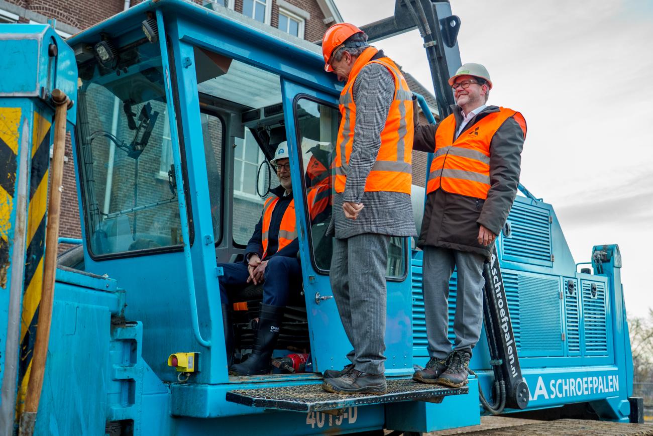 Mannen met oranje veiligheidsvestjes aan en veiligheidshelmen op bij een machine voor de start van de bouw van het kazernekwartier