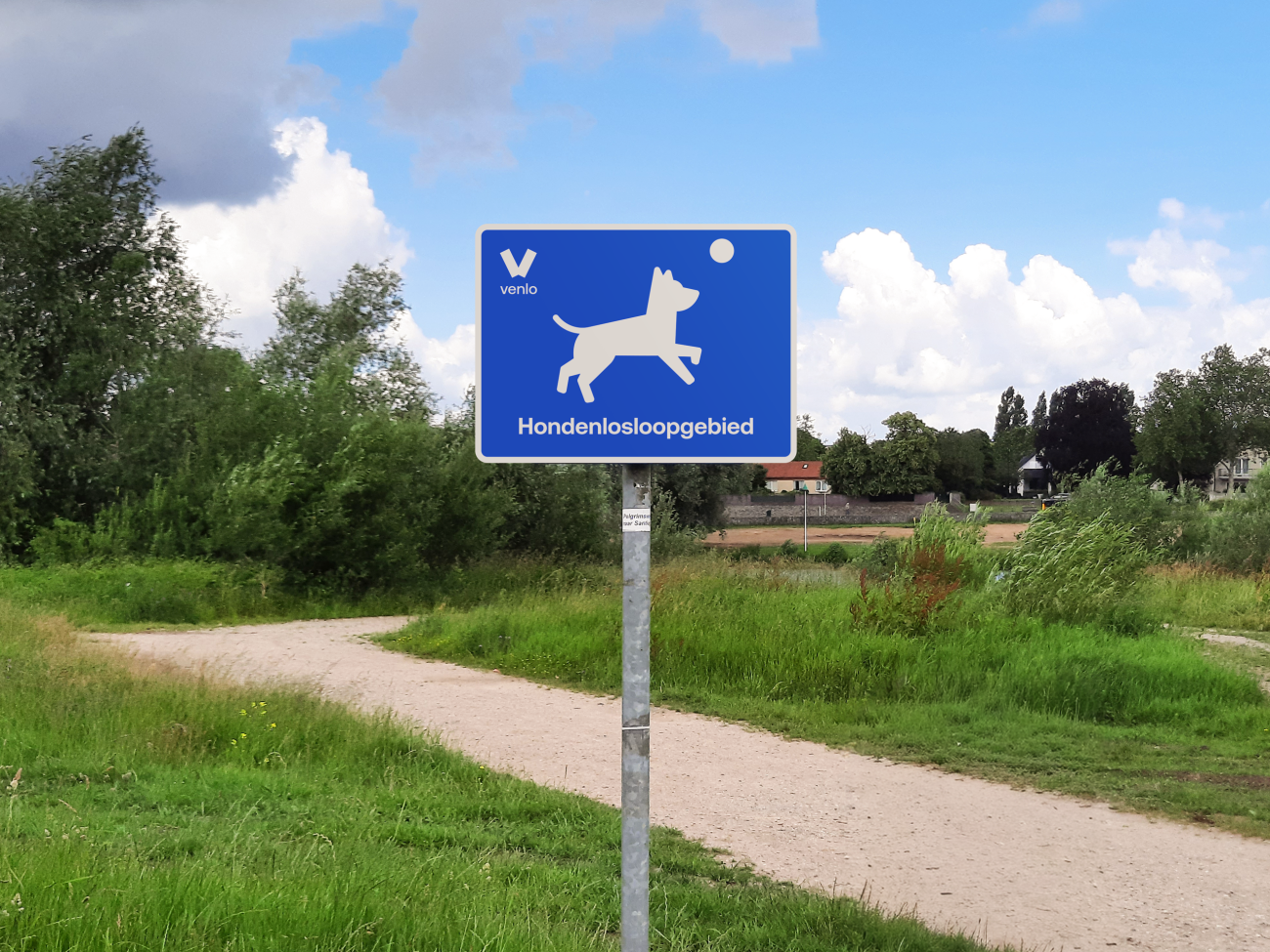 Blauw bord van de gemeente Venlo waarop een hondenlosloopgebied wordt aangegeven, geplaatst langs een wandelpad in een groen natuurgebied met gras, bomen en struiken.