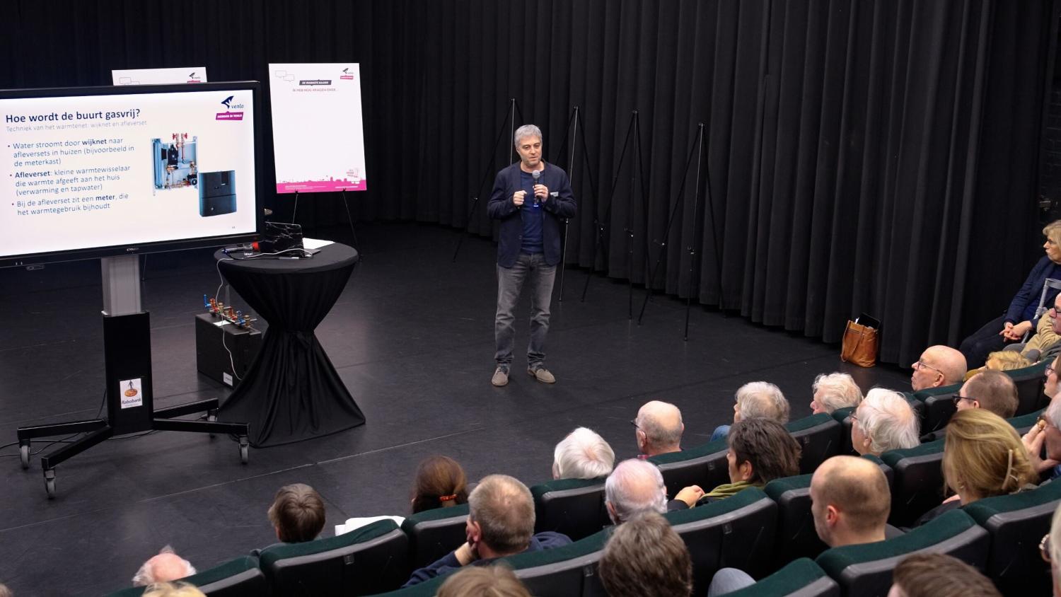 Een informatieavond over het warmtenet in Theater de Garage in Venlo, waarbij het publiek luistert naar een presentatie over het warmtenet.