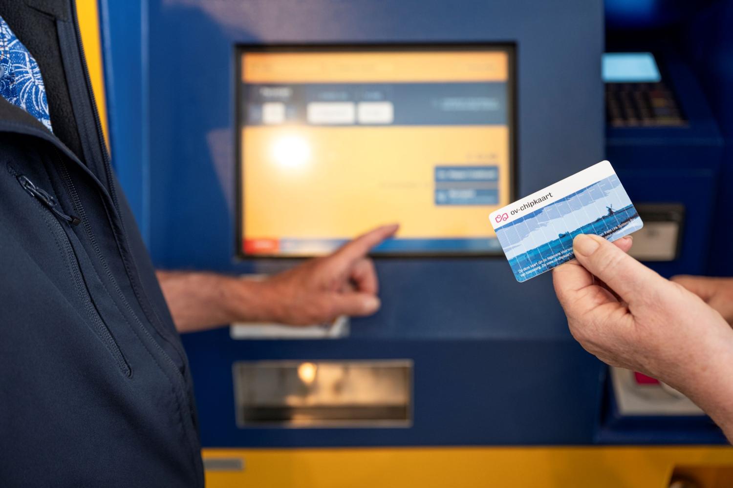 Iemand die een ov-chipkaart in de hand heeft en kijkt naar een ticket-automaat van de NS.