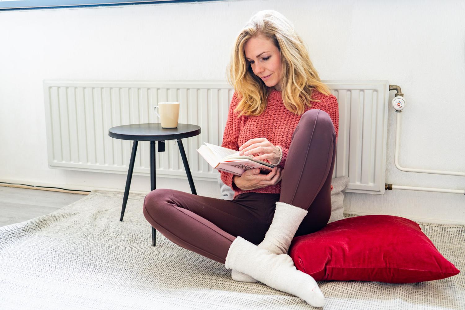 Mevrouw die een boek zit te lezen tegen de verwarming met een kop thee en dikke sokken aan
