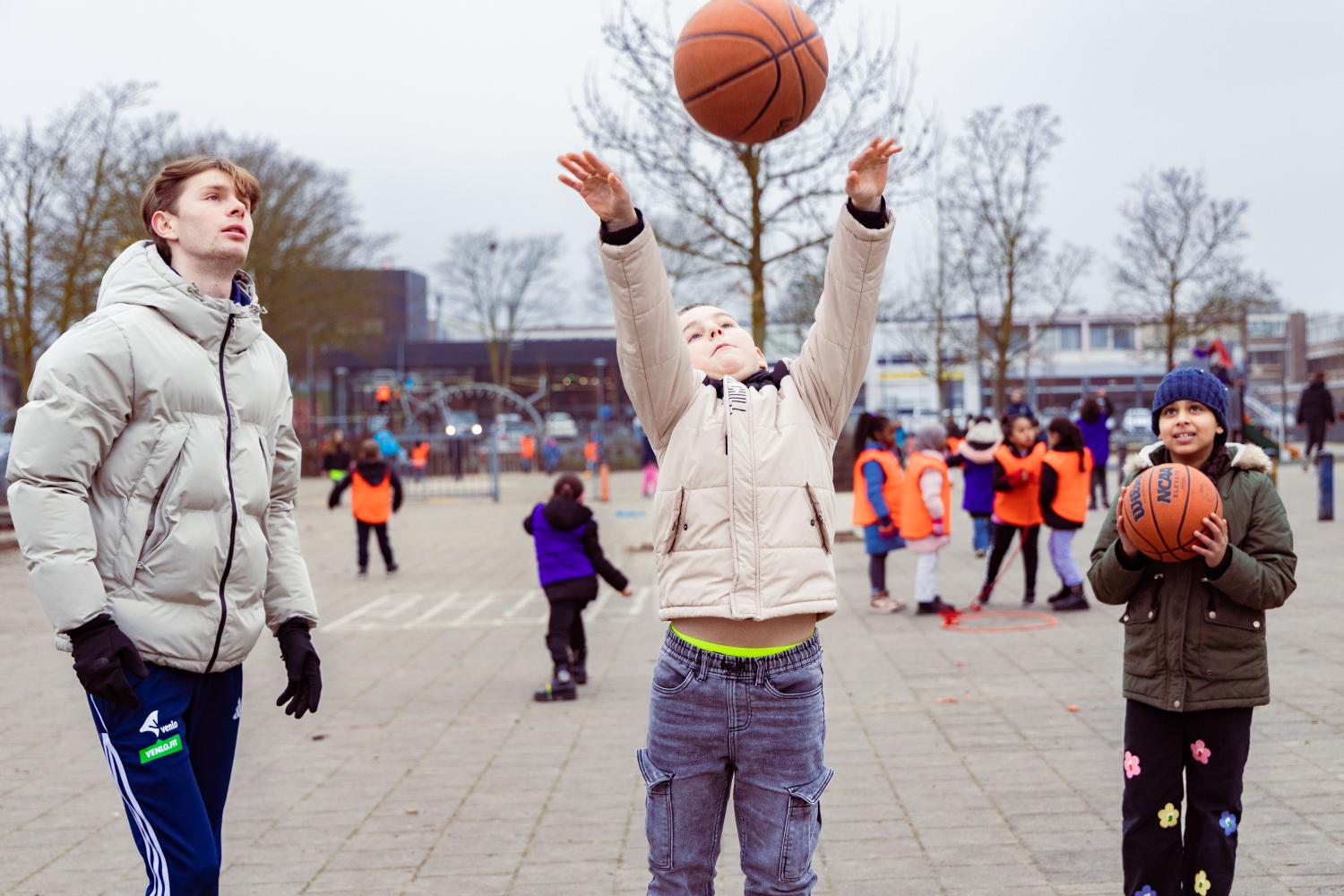 schoolplein vol kinderen, een kind gooit een basketbal in de lucht terwijl twee anderen toekijken
