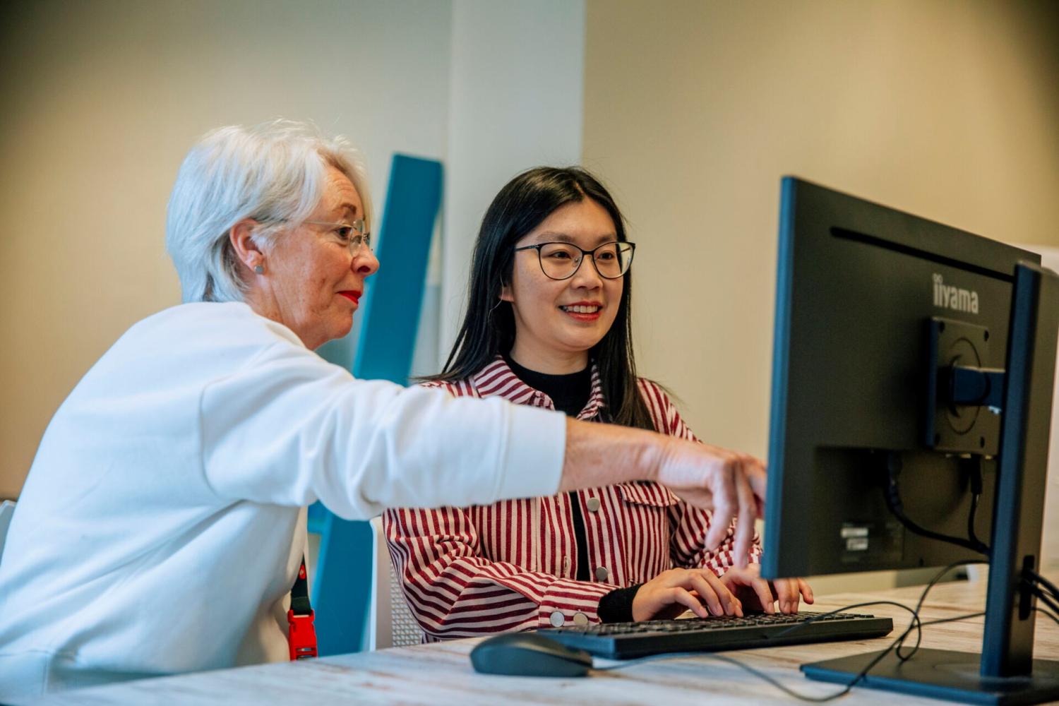 Een mevrouw die een dame uitleg geeft achter de computer