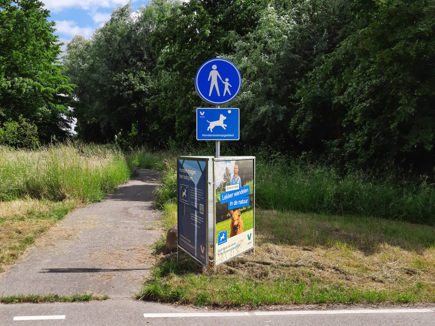Verhard wandelpad langs een bosrijke omgeving met een verkeersbord voor voetgangers en een bord dat een hondenlosloopgebied van de gemeente Venlo aangeeft. Naast het pad staat een informatiezuil over wandelen met honden in de natuur.