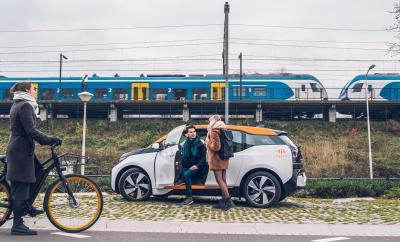 iemand die instapt in een elektrische deelauto, met een trein en fietser op de achtergrond.